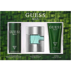 Guess Man fragrance gift set featuring a silver spray bottle with a light blue-green liquid, a dark green body spray canister, and a matching dark green body wash tube, presented on a green patterned background.