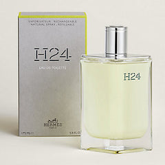 Hermès H24 Eau de Toilette perfume bottle with a silver cap next to its gray packaging box, on a light beige background.